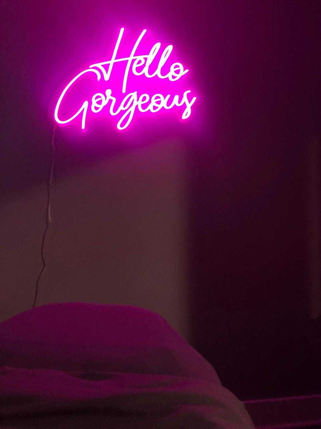 Pink neon sign reading "Hello Gorgeous" on a dark wall above a bed.