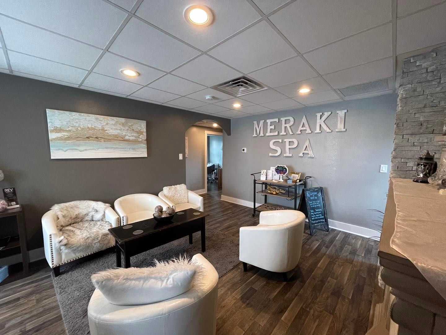 Modern spa lobby with white chairs, a coffee table, wall art, and "Meraki Spa" sign on the wall.