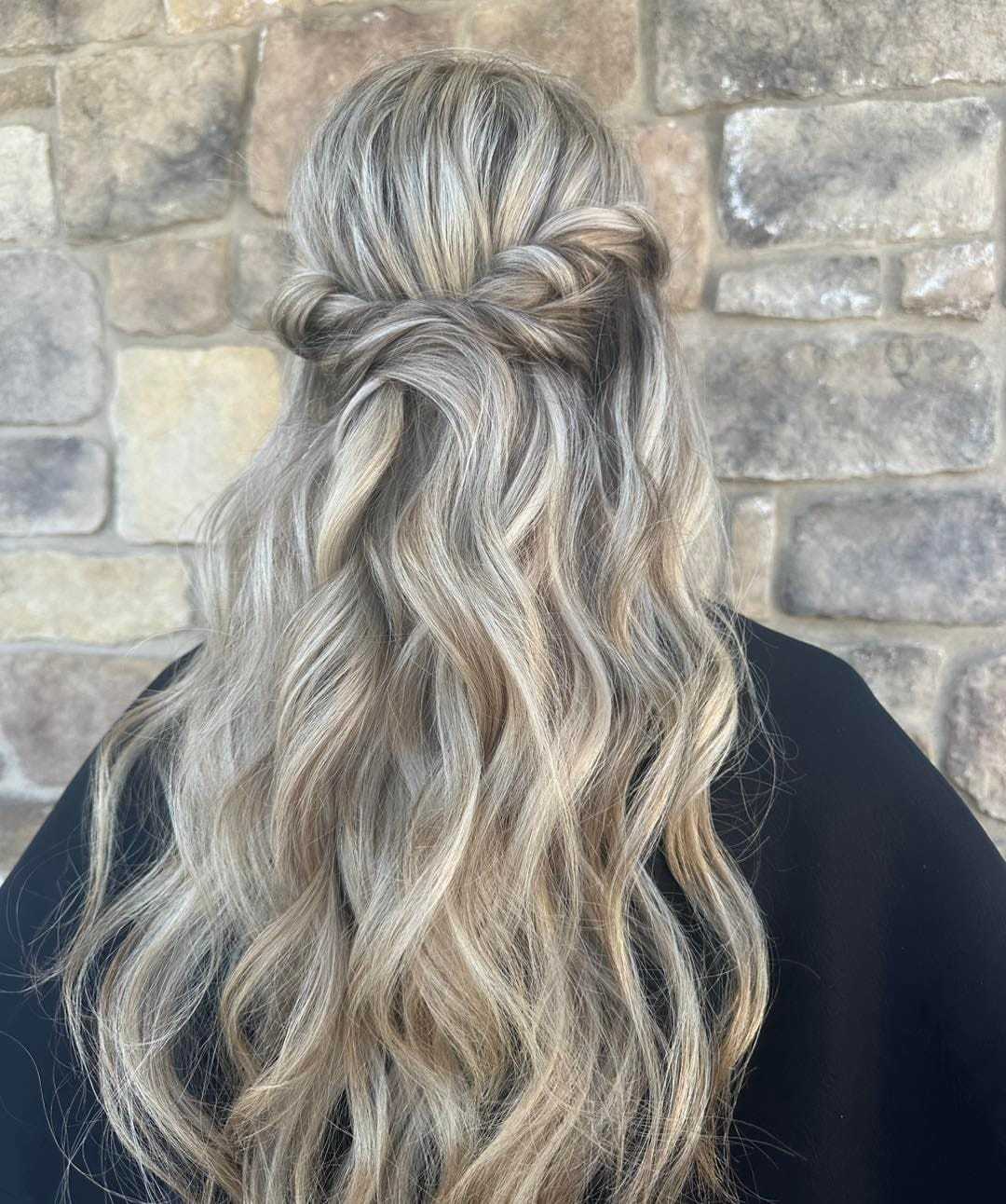 Blonde wavy hair styled in a half-up twist against a brick wall backdrop.
