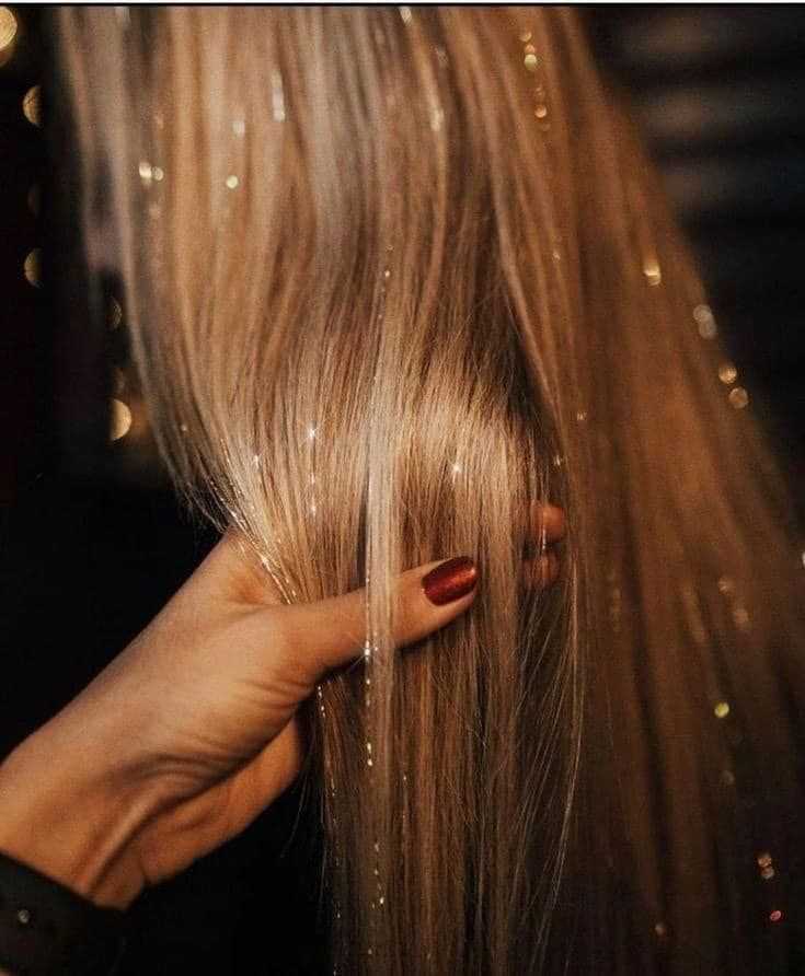 Hand with red nails combing long, shiny, blonde hair with glitter strands in a dimly lit setting.