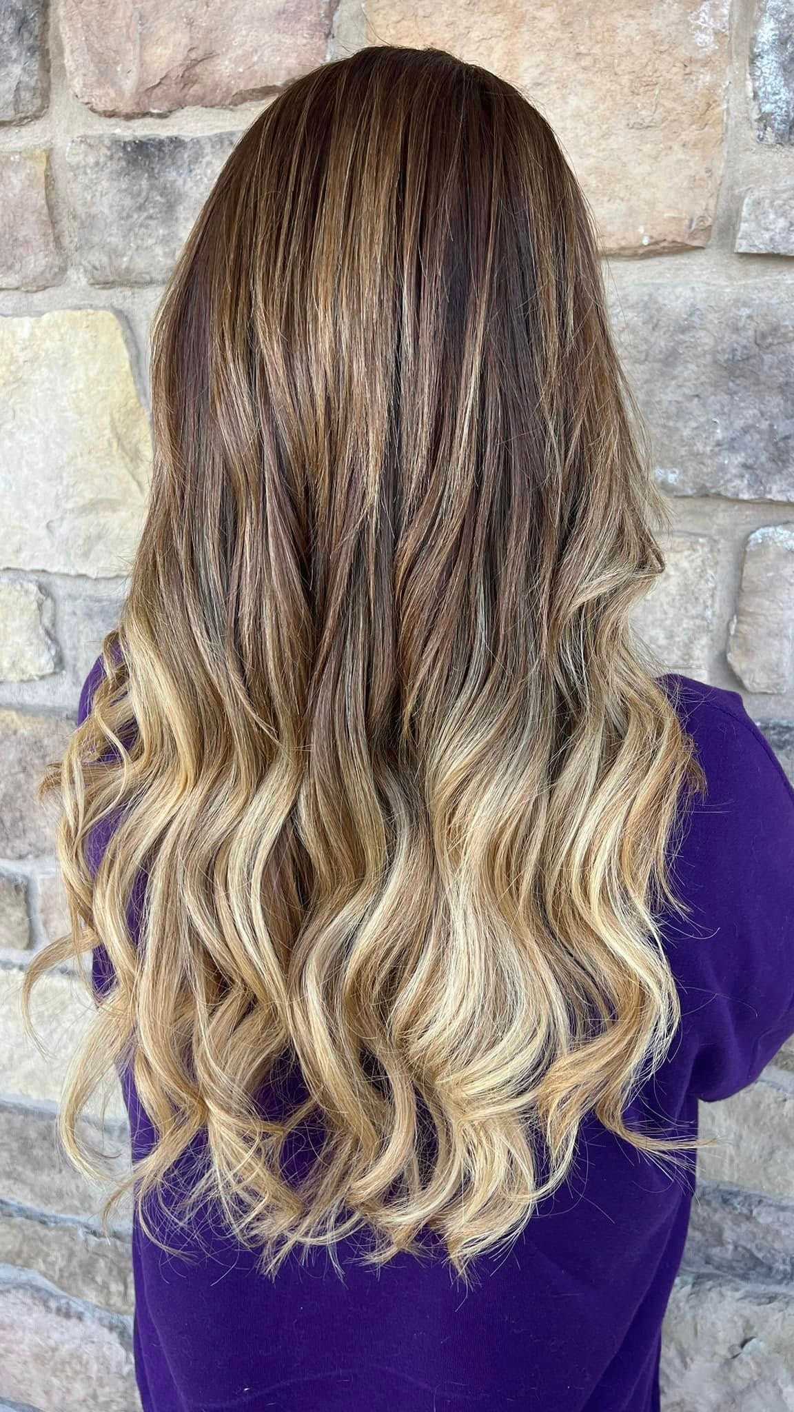 Woman with wavy, blonde ombre hair stands against a stone wall wearing a purple shirt.